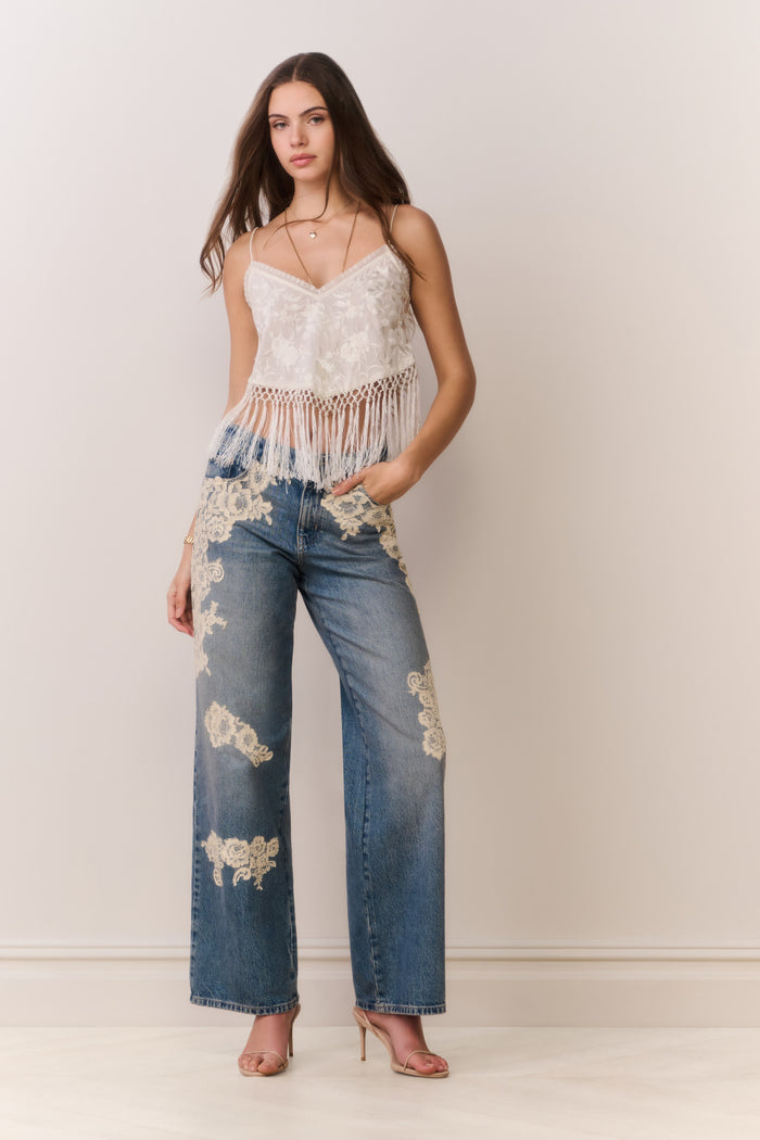 Woman wearing a white lace top with fringe and blue jeans on a plain background