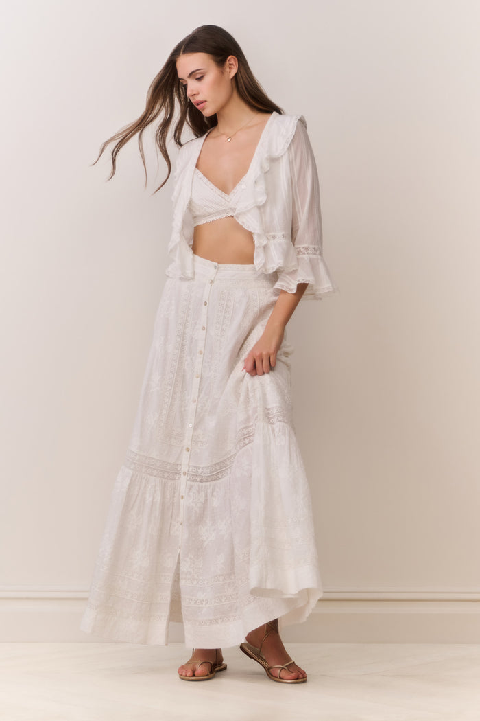 Woman wearing a white button down blouse with ruffled details and bralette top with matching maxi skirt on a plain background