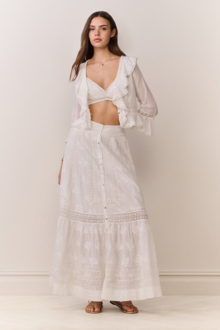 Woman wearing a white button down blouse with ruffled details and bralette top with matching maxi skirt on a plain background
