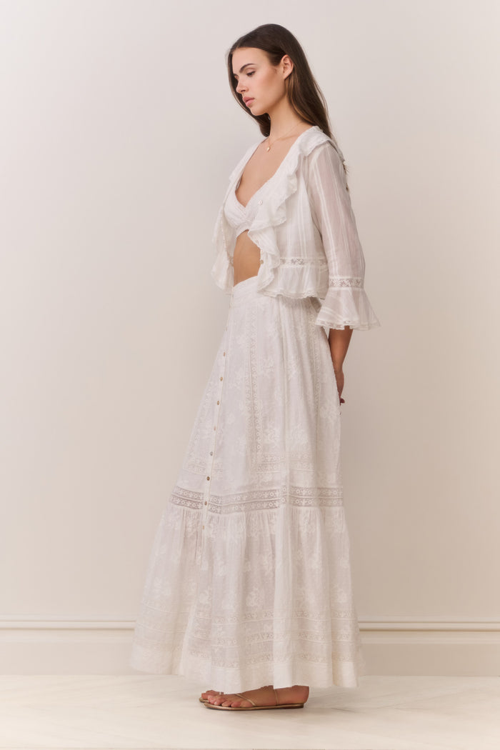 Woman wearing a white button down blouse with ruffled details and bralette top with matching maxi skirt on a plain background