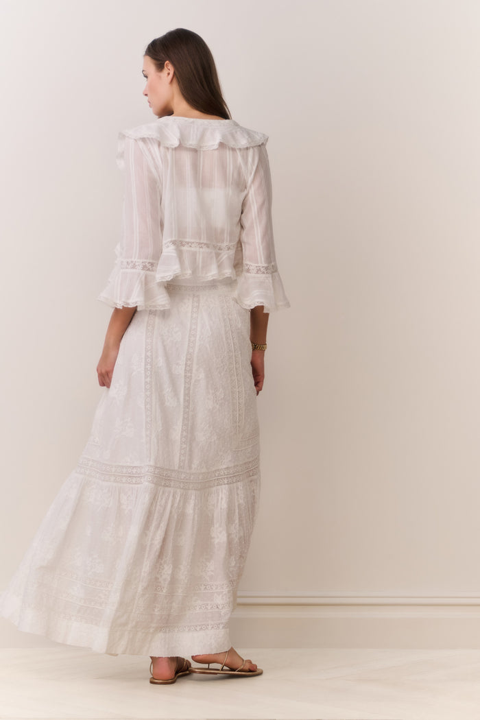 Woman wearing a white button down blouse with ruffled details and bralette top with matching maxi skirt on a plain background