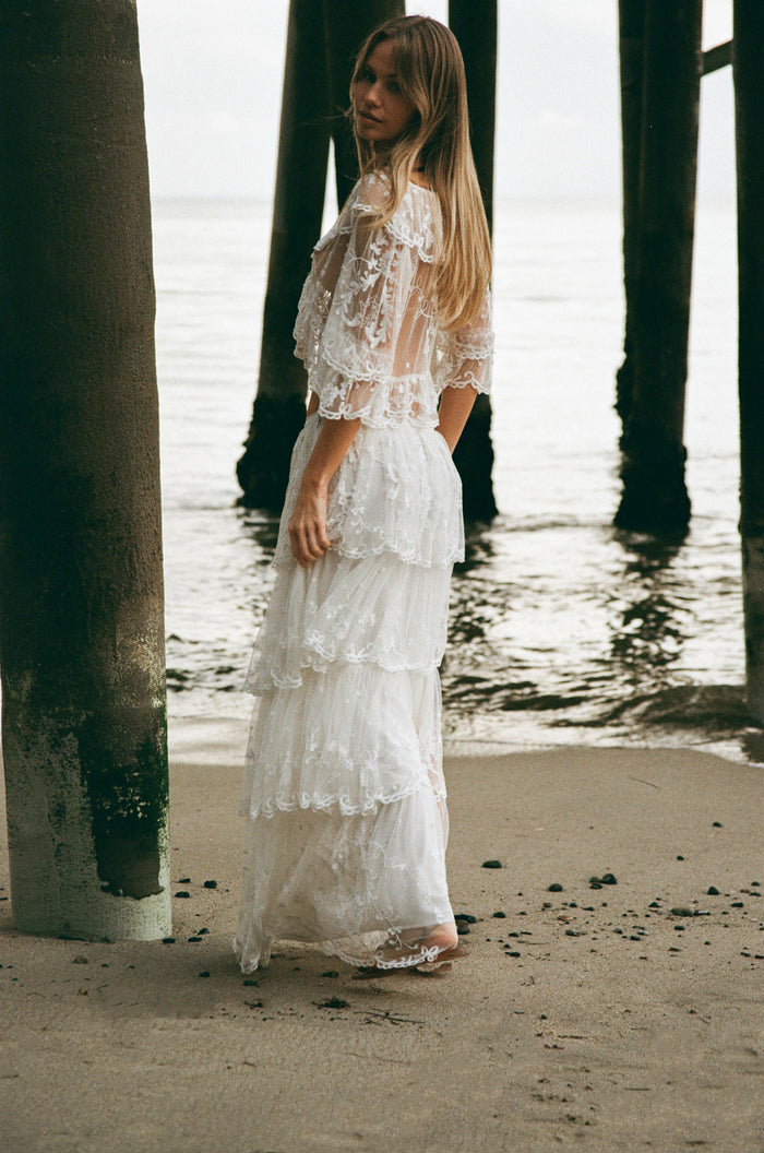 Woman wearing a white lace top and matching tiered skirt against a plain background