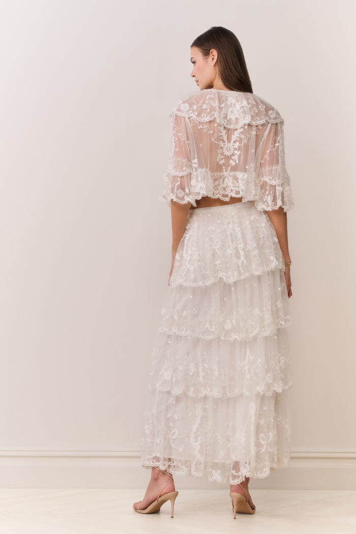 Woman wearing a white lace top and matching tiered skirt against a plain background