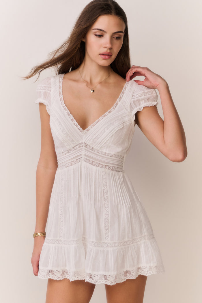 Woman wearing a white lace dress with a heart-shaped necklace against a plain background