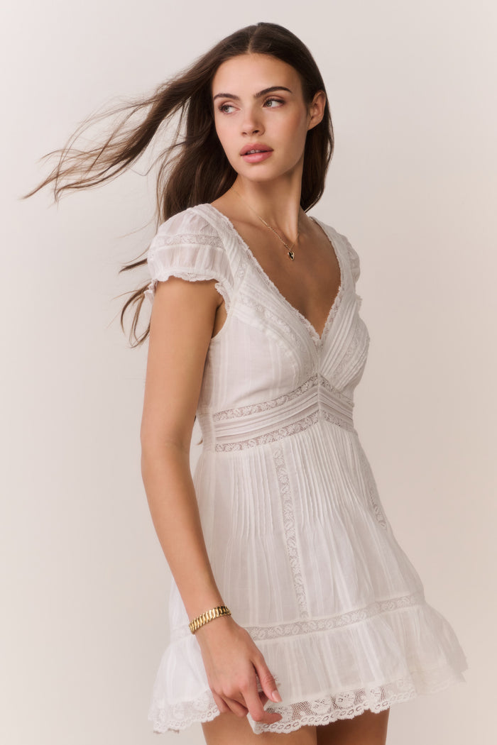 Woman wearing a white lace dress with a heart-shaped necklace against a plain background