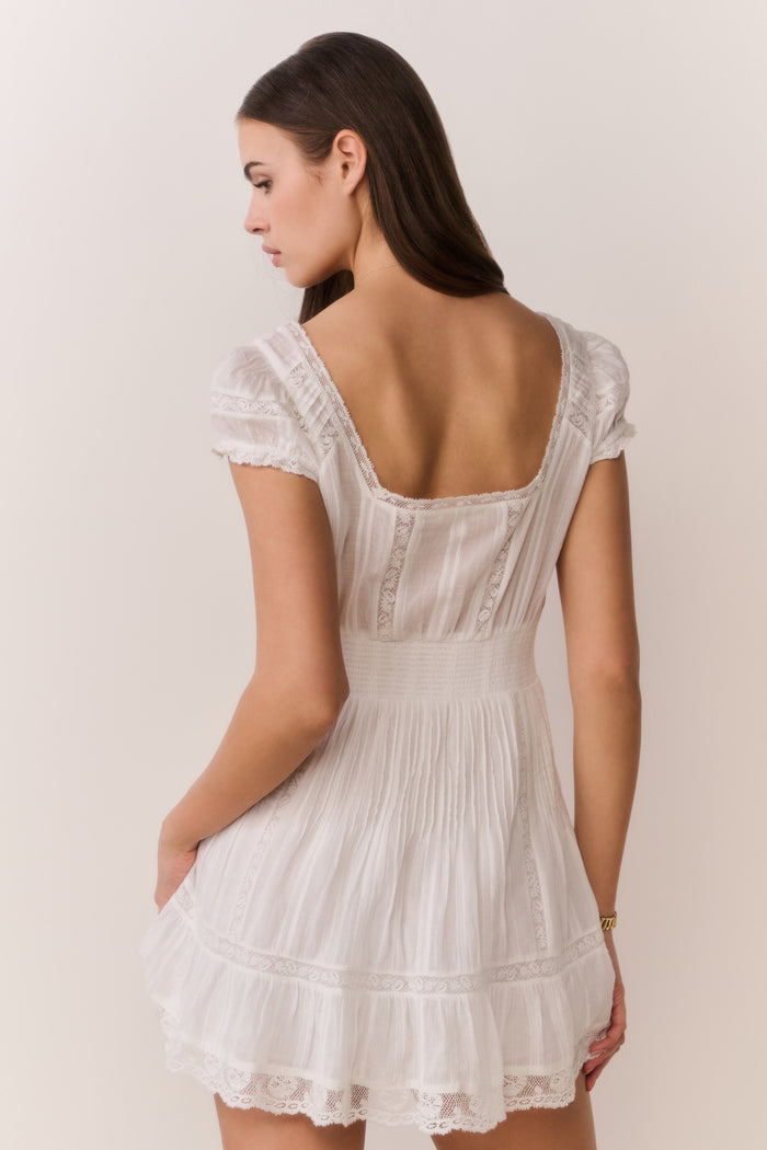 Woman wearing a white lace dress with a heart-shaped necklace against a plain background