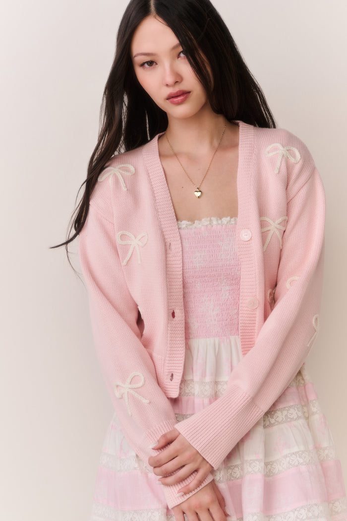 Woman wearing a pink cardigan with bow details and a pink and white lace trim skirt on a plain background