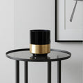 Black and gold cylindrical vase on a round glass table against a light gray wall.