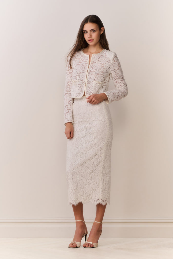 Woman wearing a white lace jacket and skirt outfit on a plain background