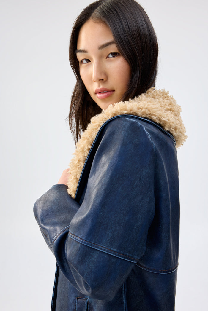 Close-up of a model wearing a navy faux leather coat with a shaggy tan faux fur collar