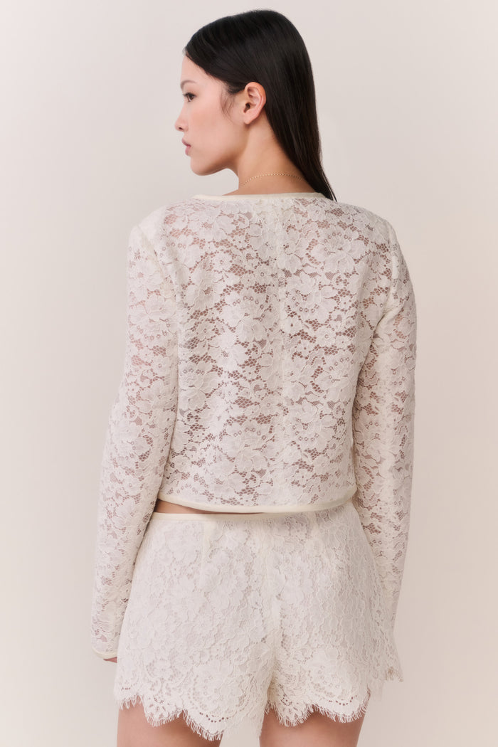 Woman wearing a white lace jacket and shorts outfit on a plain background