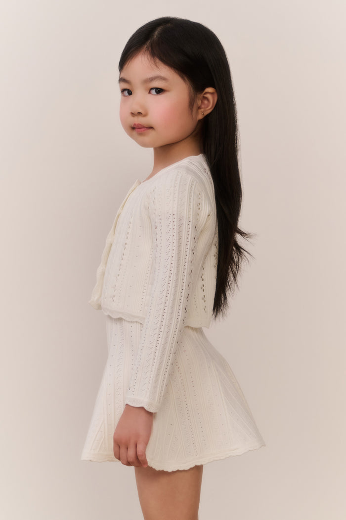 Young girl wearing a cream-colored knit cardigan and skirt outfit against a plain background