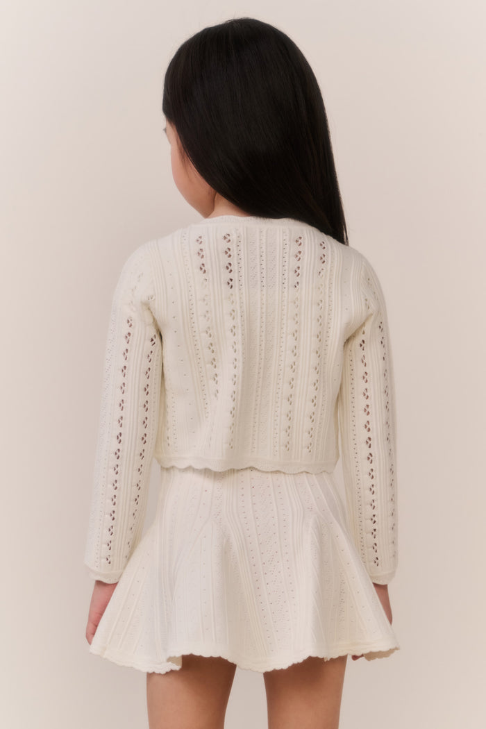 Young girl wearing a cream-colored knit cardigan and skirt outfit against a plain background