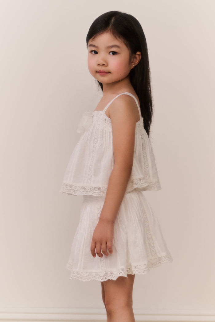 Little girl wearing a white lace trim camisole and matching skirt against a plain background