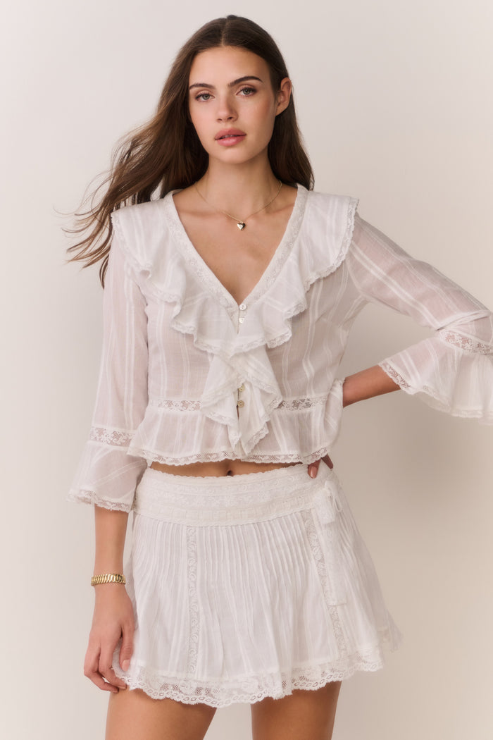 Woman wearing a white lace long sleeve blouse with ruffle trim and matching skirt against a plain background