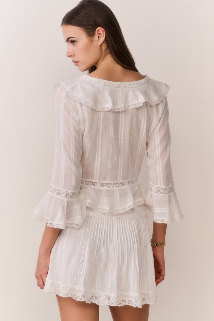 Woman wearing a white lace long sleeve blouse with ruffle trim and matching skirt against a plain background
