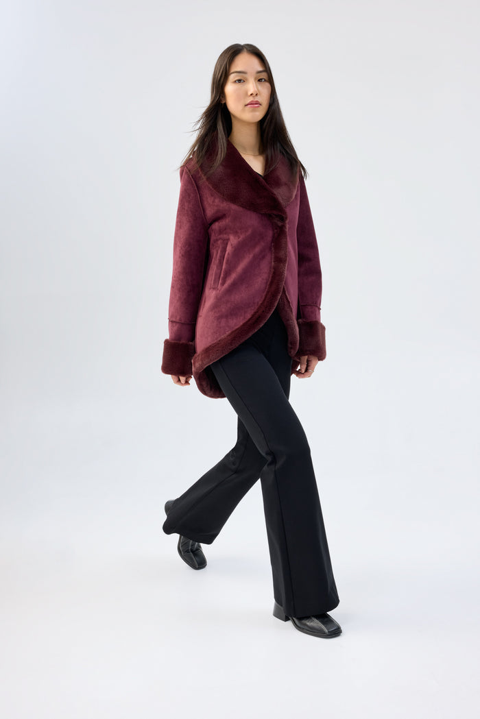 Model walking, wearing a burgundy faux fur jacket with shawl collar and wrap-front, contrast plush cuffs, over black flared trousers