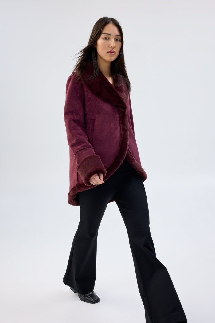 Model wearing a burgundy faux fur jacket with shawl collar and wrap-front, contrast plush cuffs, worn over black flared trousers