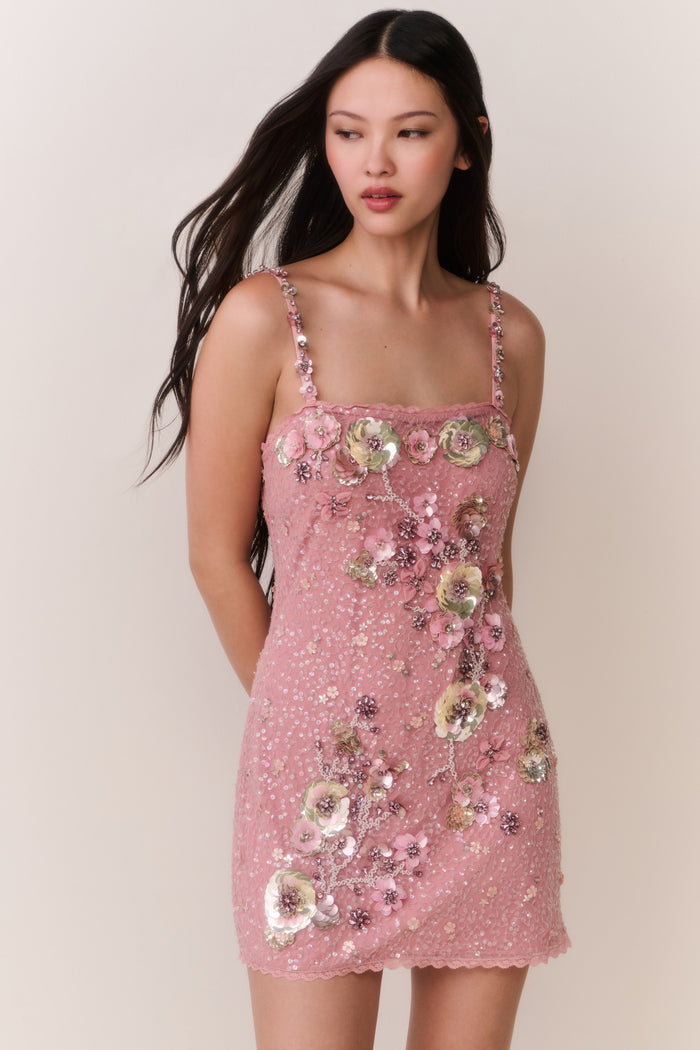 Woman wearing a pink dress with floral embellishments against a plain background