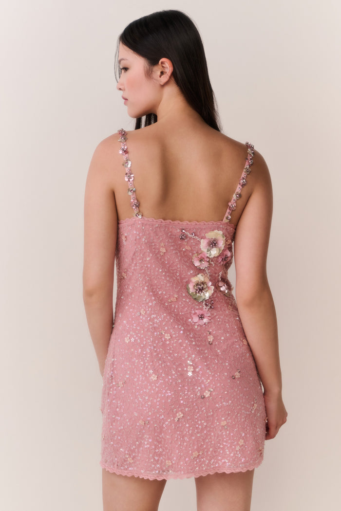 Woman wearing a pink dress with floral embellishments against a plain background