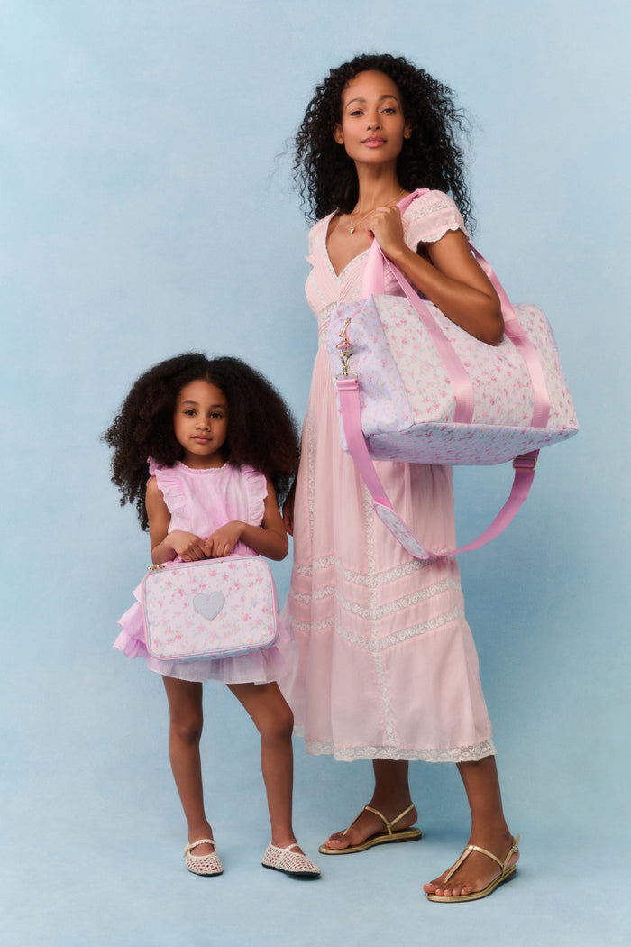 Two women, one adult and one child, standing together holding matching pink bags against a light blue background.