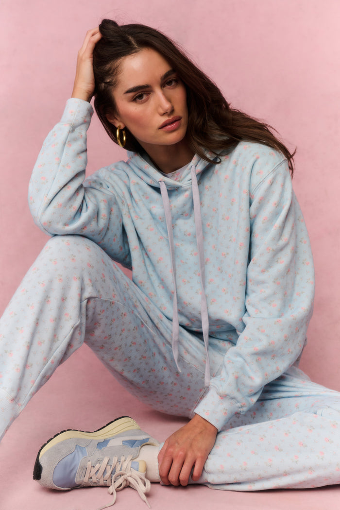 Woman wearing a light blue floral hoodie and pants sitting on a pink background