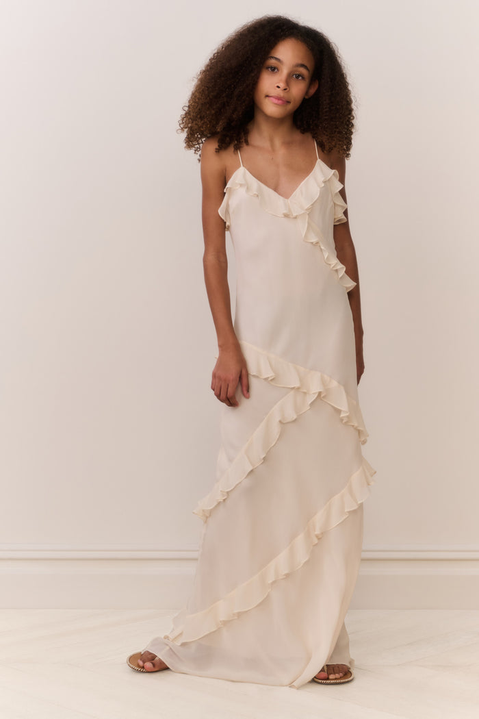 Tween girl wearing a flowing white sleeveless dress with ruffles on a plain background