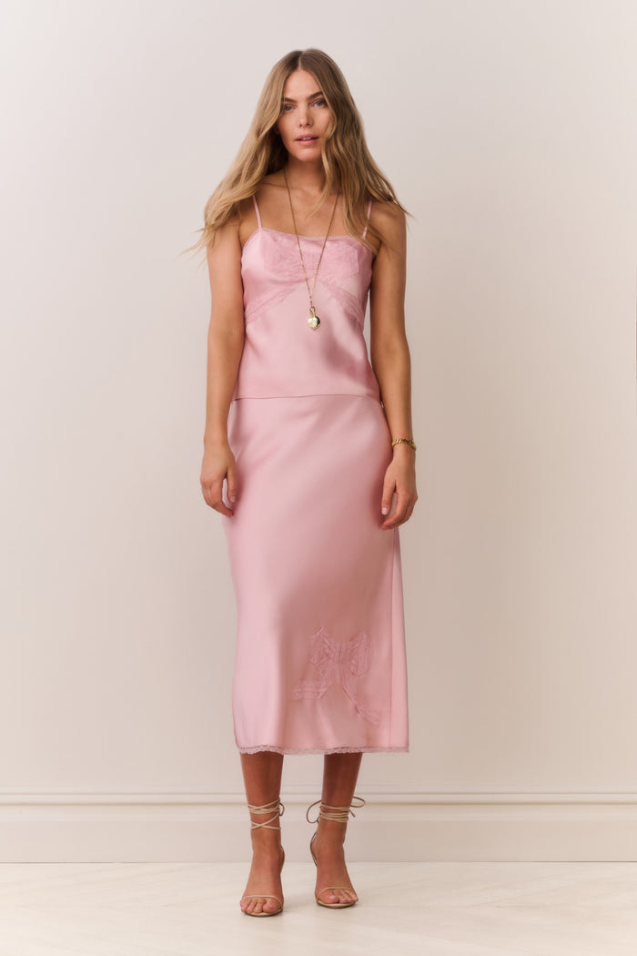 Woman wearing a pink satin cami and matching skirt with thin straps and bow detail on a plain background