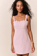 Woman wearing a pink dress with thin straps and scalloped neckline against a beige background